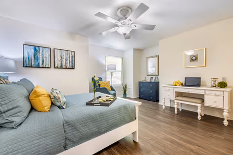 A bright and spacious bedroom with a large bed covered in teal bedding and yellow accent pillows. There is a white ceiling fan above, two framed artworks on the wall, a blue armchair with a yellow pillow near a window, a dark blue dresser with a mirror above it, and a white desk with a laptop and decorative items. The floor is wood, and the walls are light-colored.