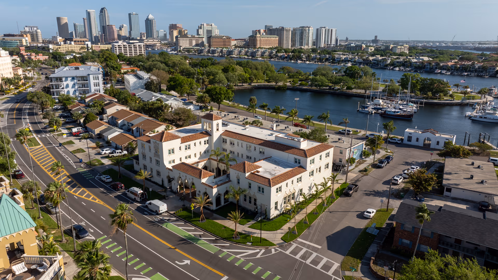 Aerial view of Hudson Manor Assisted Living, a large white building with a red-tiled roof located near a waterfront with boats docked. The surrounding area includes roads, palm trees, and other buildings, with a city skyline visible in the background under a clear blue sky.