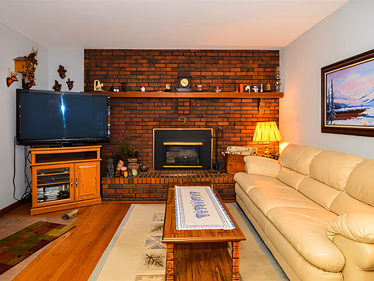 Cozy living room with a brick fireplace, cream leather sofa, TV on a wooden stand, and a wooden coffee table.