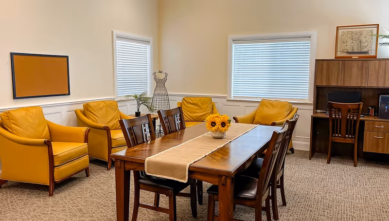 A cozy room with a wooden table and six chairs in the center, decorated with a beige table runner and a vase of sunflowers. Surrounding the table are four mustard-yellow armchairs. The room has two windows with white blinds, a corkboard on the wall, a decorative wire dress form, a small plant, and a wooden desk with a chair and framed artwork on top.