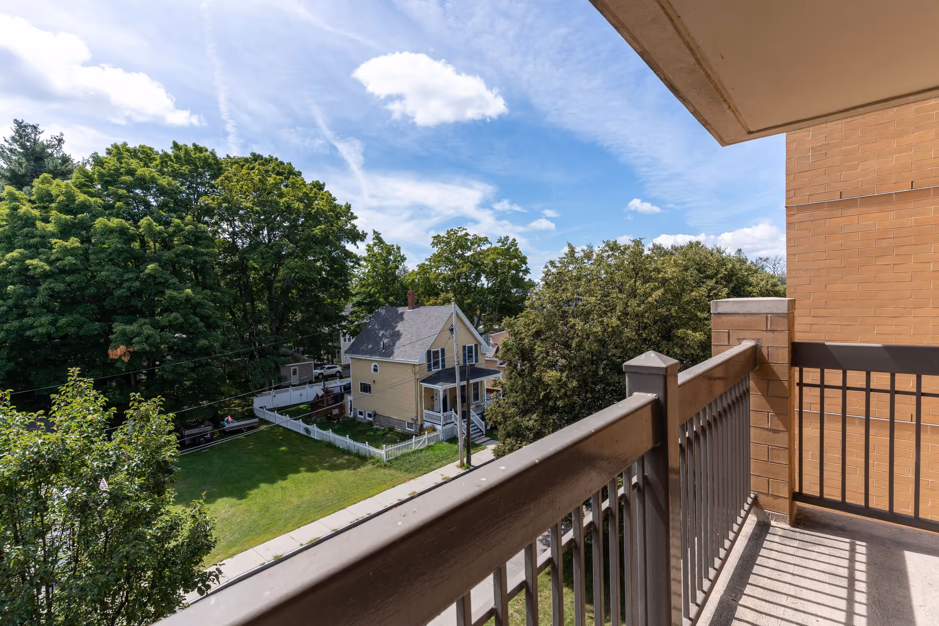 View from a balcony overlooking a tree-lined neighborhood with a yellow house and grassy yard.