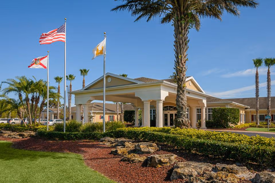 Front entrance of a single-story care facility with a covered porte-cochère, palm trees, and three flagpoles flying flags.