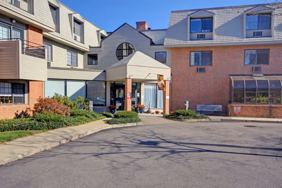 Exterior view of a senior living facility building with a combination of brick and light-colored siding. The entrance has a covered porch with a small garden area and potted plants near the door. There is a paved driveway and sidewalk leading to the entrance, with some bushes and greenery along the building.