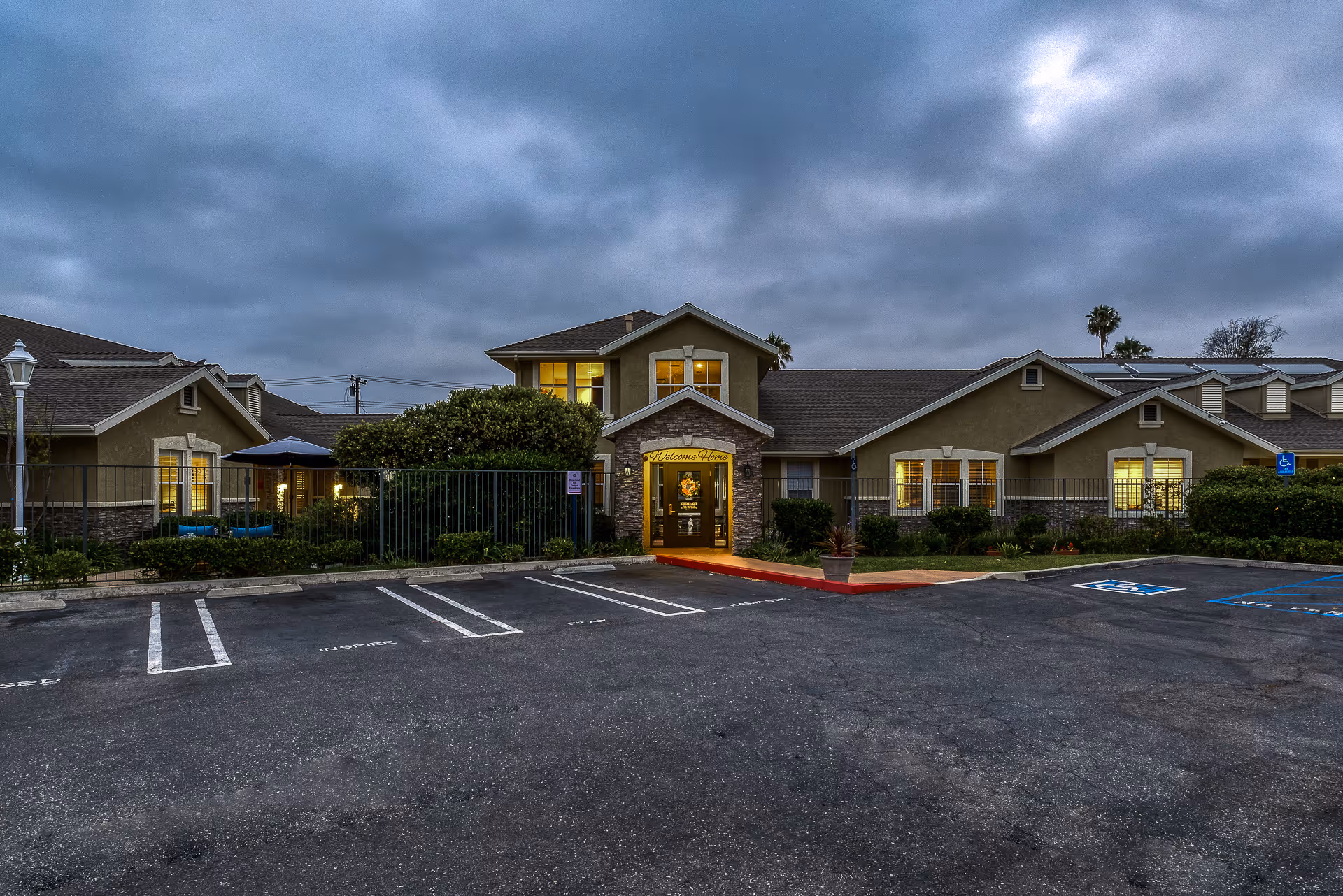 Front entrance of a single-story senior living building at dusk with lit windows and empty parking spaces.
