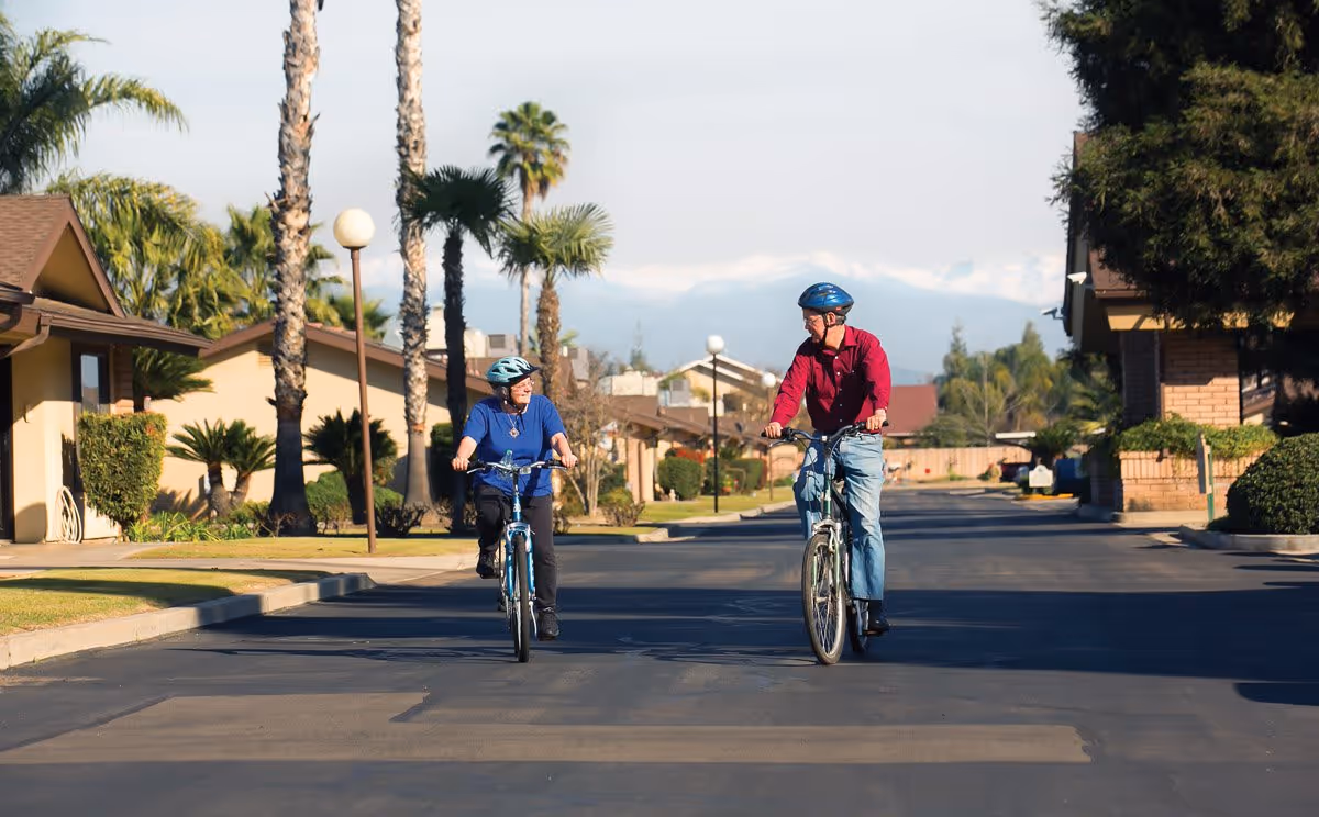 Two elderly people wearing helmets riding bicycles on a paved street in a residential area with palm trees and houses on either side under a clear sky.