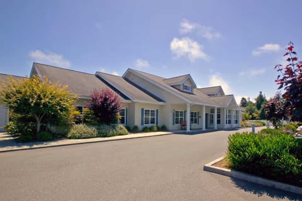 Exterior view of a single-story building with a sloped roof, white siding, and multiple windows. The building is surrounded by landscaped greenery including bushes and trees under a clear blue sky.