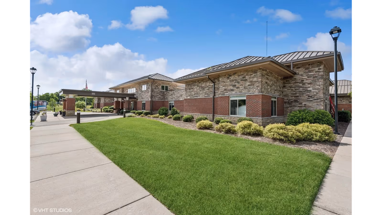 Exterior front entrance of a single-story brick and stone senior living facility with a covered drive, manicured lawn, and walkway under a blue sky.