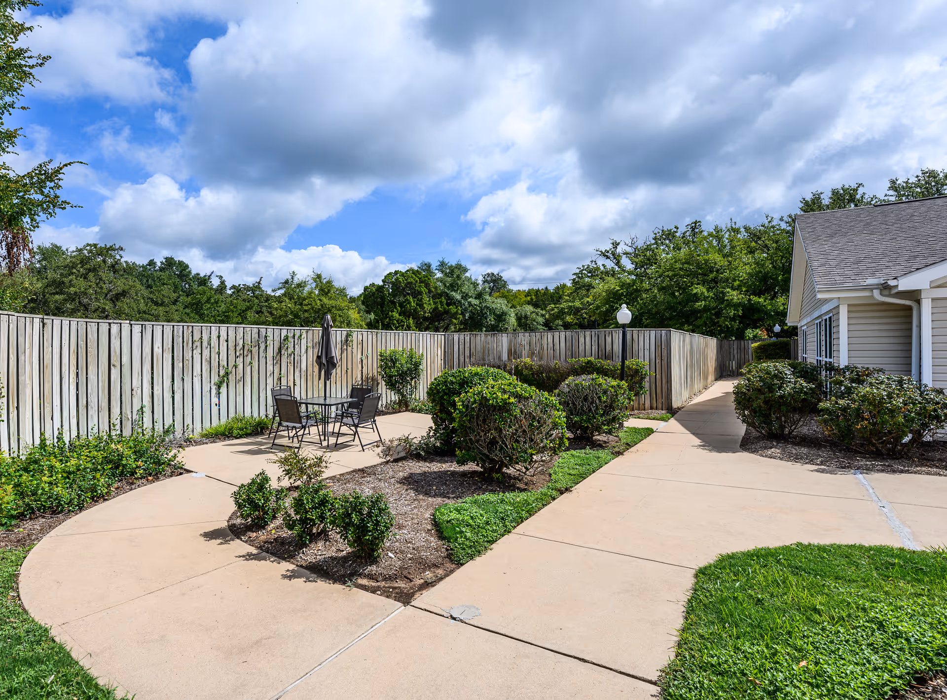 Outdoor patio area with a round table and four chairs, surrounded by a wooden fence and landscaped bushes. A concrete pathway leads past the patio alongside a building with beige siding under a partly cloudy sky.