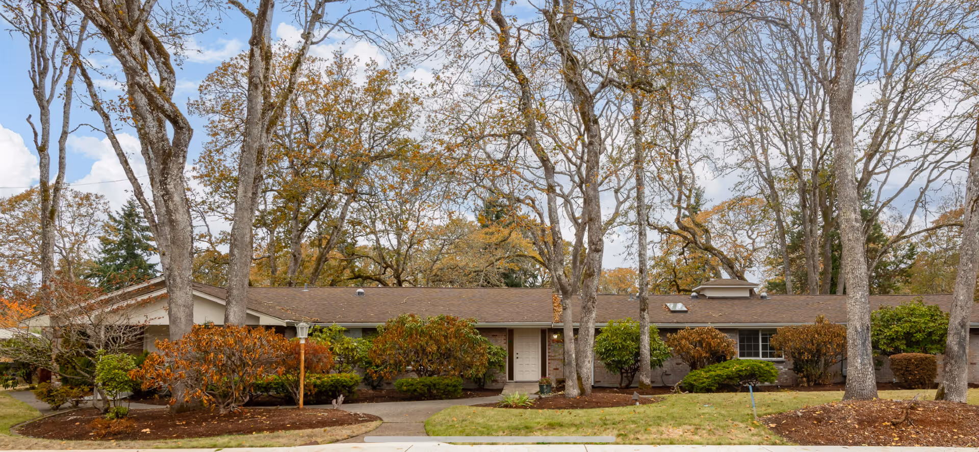 Single-story residential building with a brown roof surrounded by leafless trees and various shrubs in front, under a partly cloudy sky.