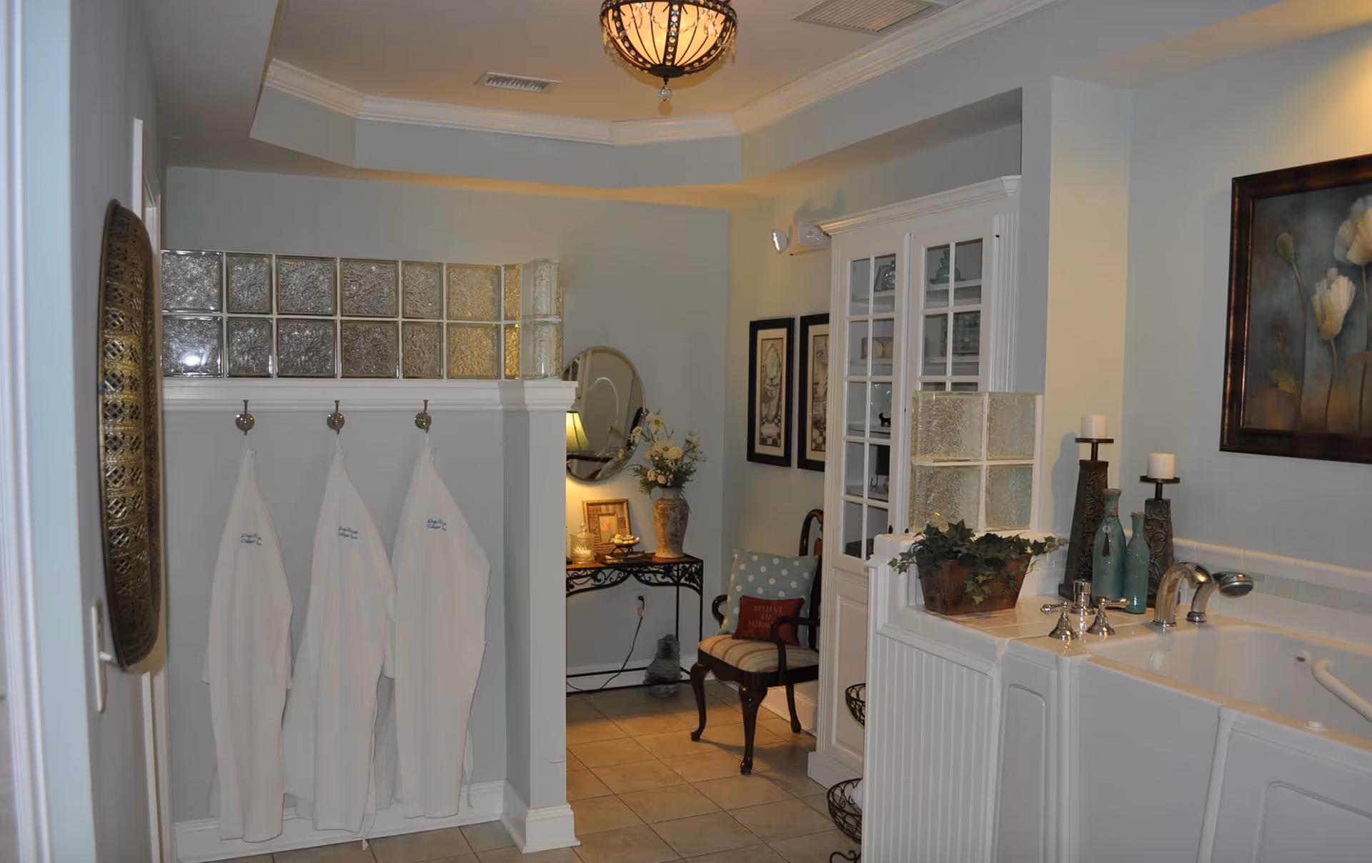 Interior view of a bathroom area in an assisted living facility featuring three white robes hanging on hooks on a wall with glass block accents. To the right is a walk-in bathtub with decorative candles, vases, and a framed floral painting above it. In the background, there is a small seating area with a wooden chair with a cushion and a pillow that says 'Believe in Miracles,' a round mirror, a table with a lamp, flowers, and framed pictures on the wall.