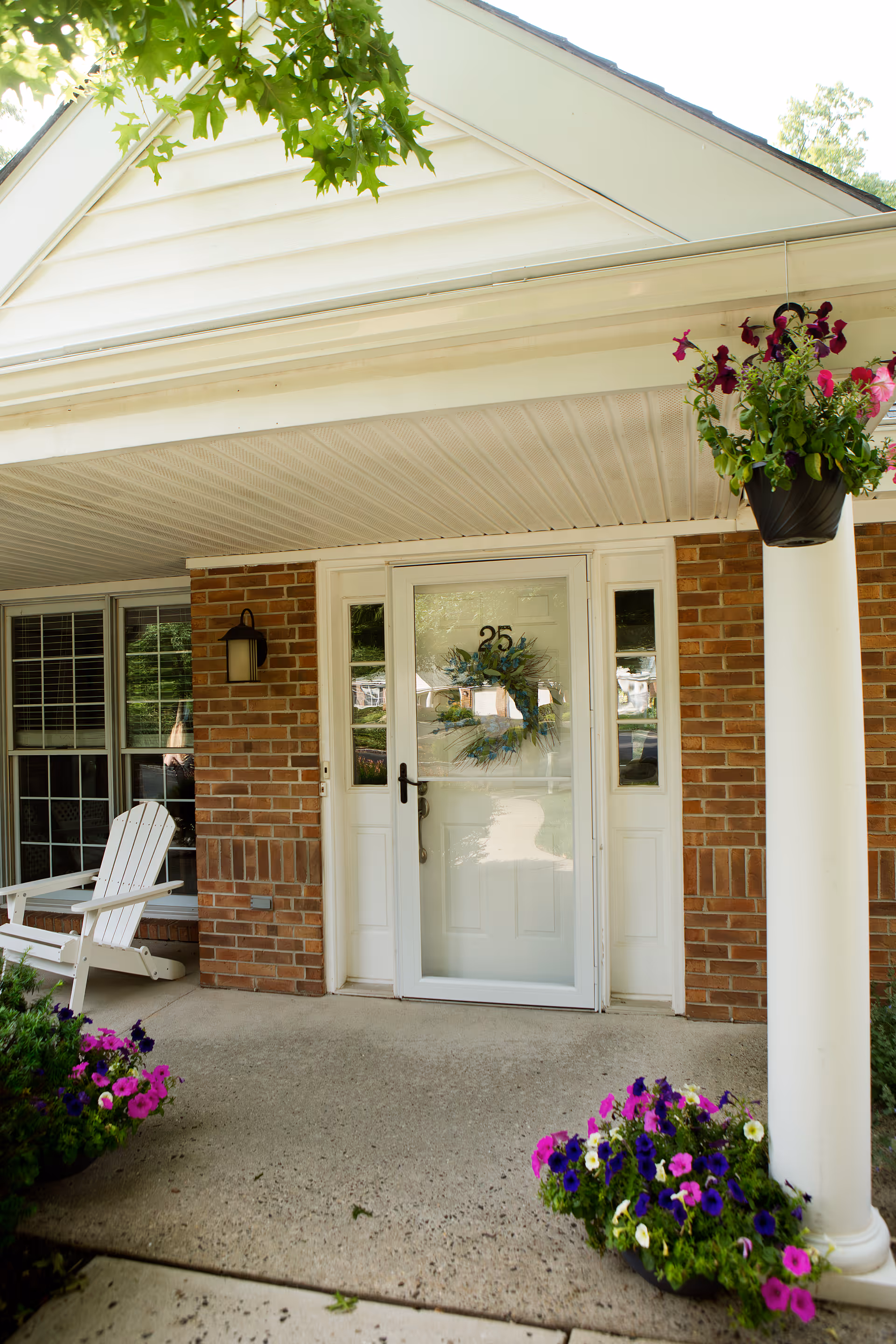 Front porch entrance of a brick building with a glass door numbered 25, hanging and potted flowers, a white column, and an Adirondack chair.