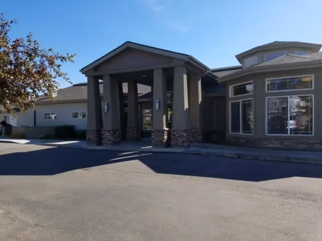 Exterior view of a single-story building with a covered entrance supported by four columns with stone bases. The building has large windows and a clear blue sky above.