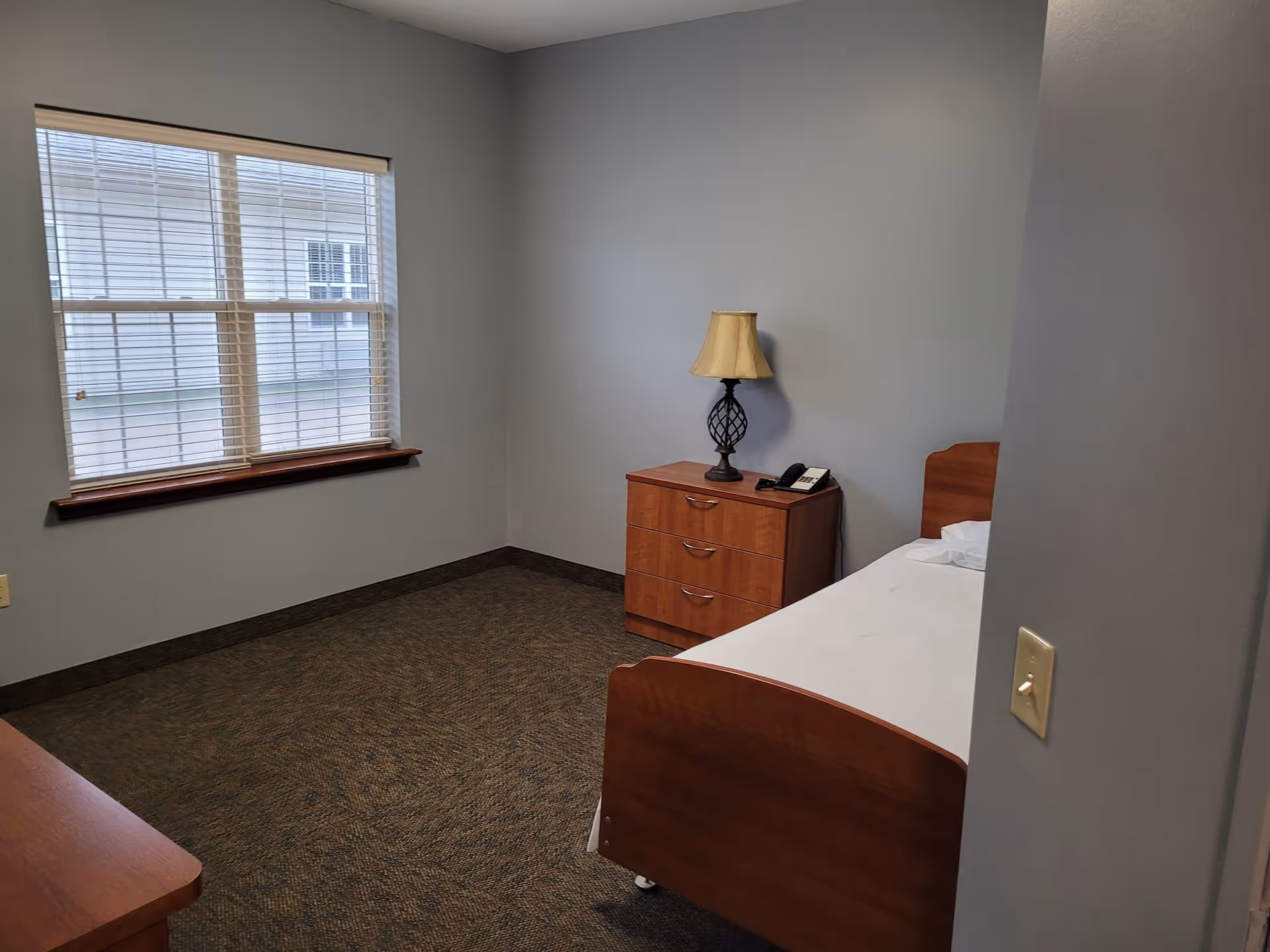 A simple bedroom with a single wooden bed with white bedding, a wooden dresser with a lamp and a telephone on top, a window with blinds, and gray walls.