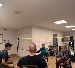 Four elderly individuals seated in chairs playing a seated volleyball game indoors with a yellow net set up between them in a well-lit room with white walls and ceiling lights.
