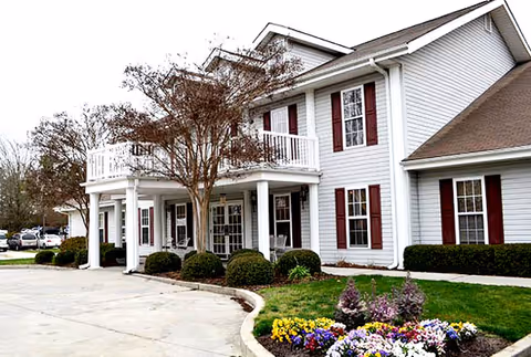 Exterior view of a two-story senior living facility building with white siding and red shutters. The entrance features a covered porch with white columns and a balcony above. There are neatly trimmed bushes and a colorful flower bed in front of the building, along with a paved driveway.
