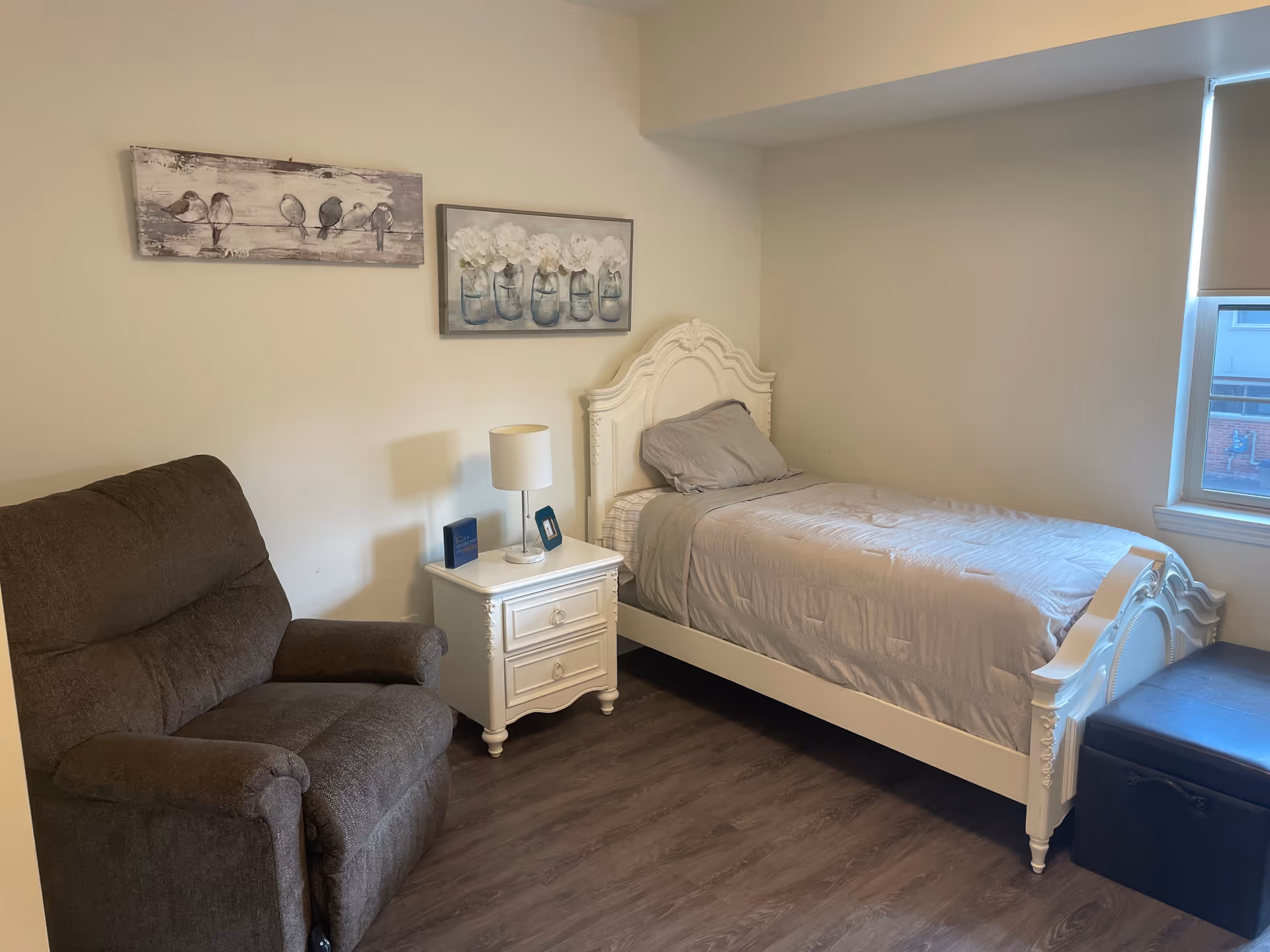 A small bedroom with a single white ornate bed with gray bedding, a white nightstand with a lamp and small framed pictures, a dark brown recliner chair, two paintings on the wall, and a window with a roller shade partially down.