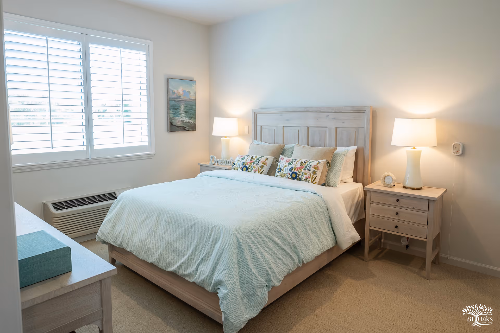 A bright and cozy bedroom with a large bed featuring a light blue bedspread and multiple decorative pillows. There are two wooden nightstands on either side of the bed, each with a white lamp. A window with white shutters lets in natural light, and a painting of a seascape hangs on the wall beside the bed. The room has light-colored walls and carpeted flooring.