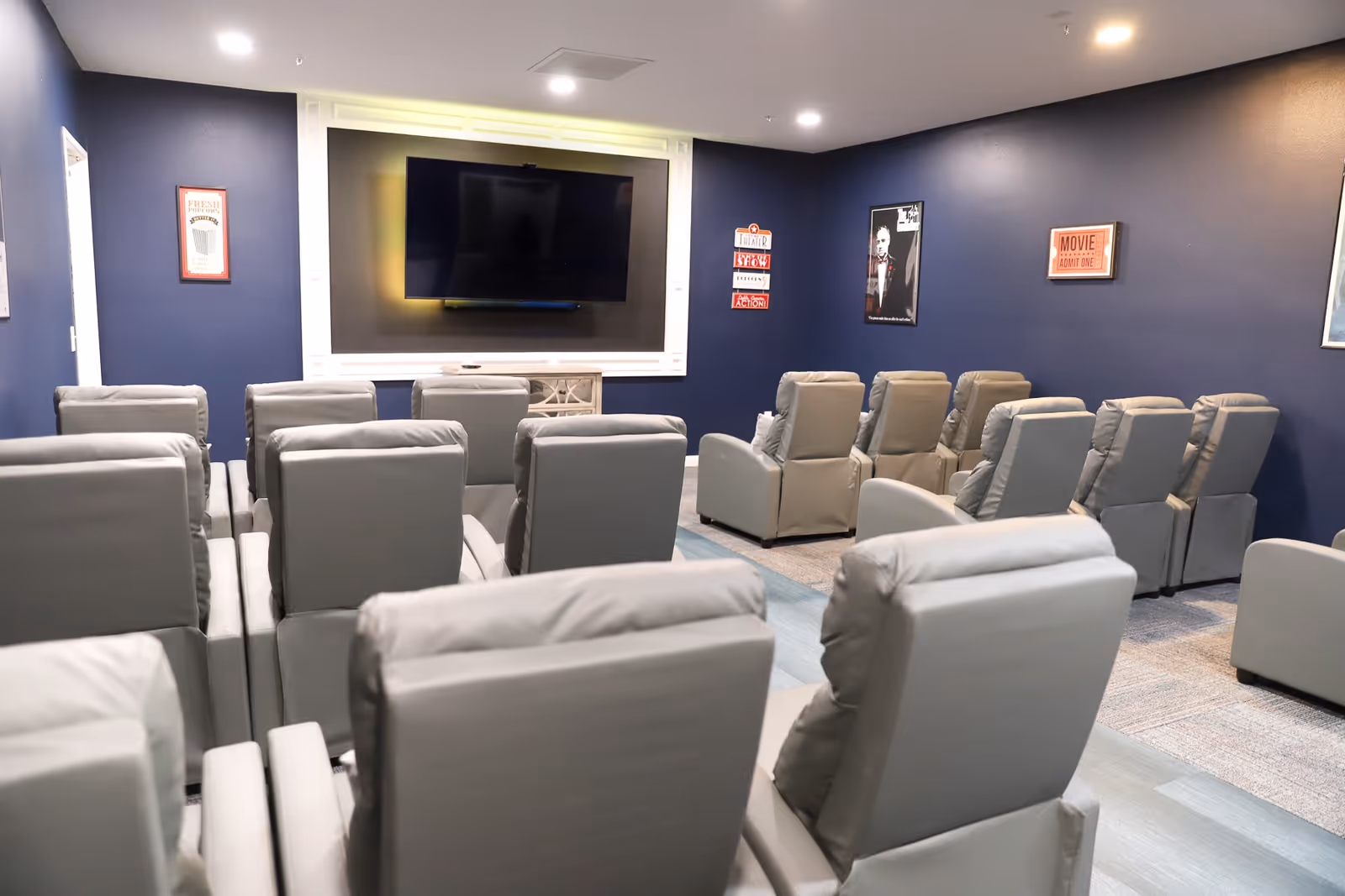 A small movie theater room with multiple rows of gray reclining chairs facing a large flat-screen TV mounted on a dark blue wall. The walls are decorated with framed movie-themed posters and signs.