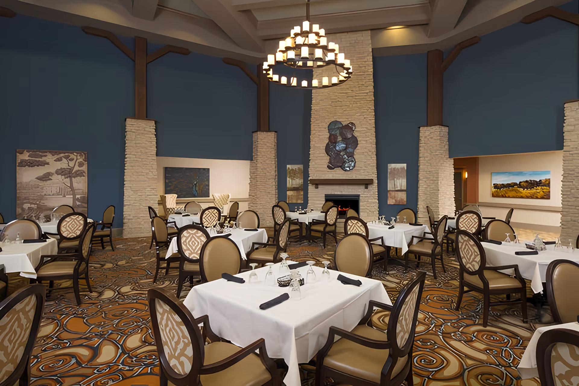 A spacious dining room with multiple tables covered in white tablecloths, each set with glasses and napkins. The room features a high ceiling with exposed beams, a large chandelier, and a stone fireplace with decorative artwork above it. The walls are painted blue and adorned with various paintings. The carpet has an intricate swirling pattern in shades of brown and orange.