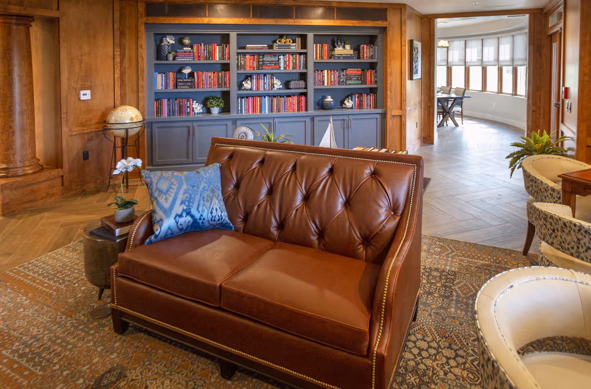 A cozy living room area featuring a brown leather tufted loveseat with a blue patterned pillow, a patterned area rug, and a built-in bookshelf filled with books and decorative items. The room has wooden paneling and columns, with a globe on a stand to the left and a curved hallway with large windows in the background.