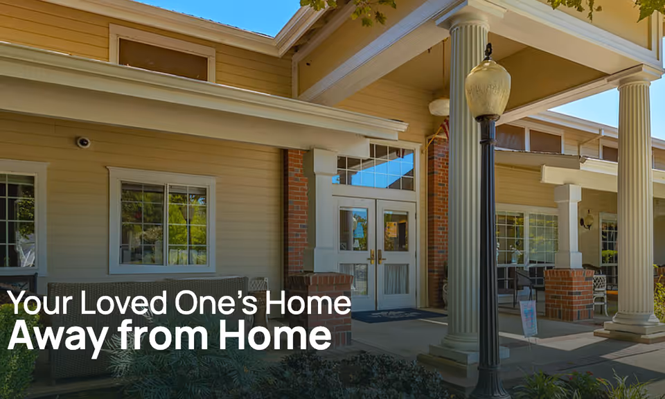 Covered entrance of a senior living facility showing columns, double glass doors, outdoor seating and a lamppost.