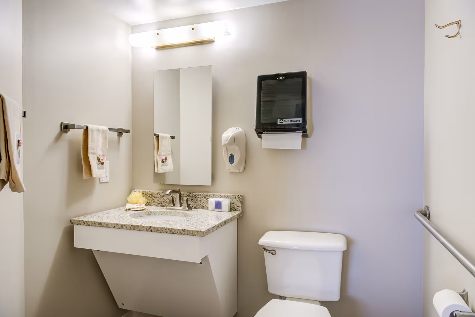A clean and simple bathroom with a white toilet and a granite countertop sink. Above the sink is a rectangular mirror with a light fixture above it. There are beige hand towels with floral embroidery hanging on a towel rack to the left, a soap dispenser and a paper towel dispenser mounted on the wall above the toilet, and a grab bar on the right wall.