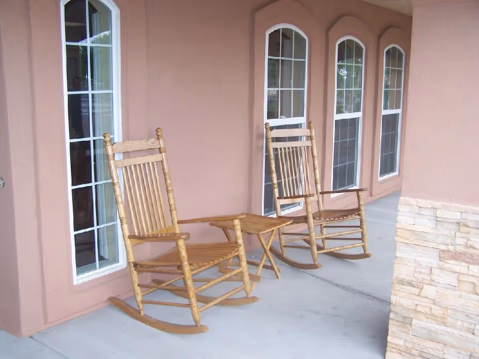 Two wooden rocking chairs and a small wooden table on a covered porch with peach-colored walls and arched windows.