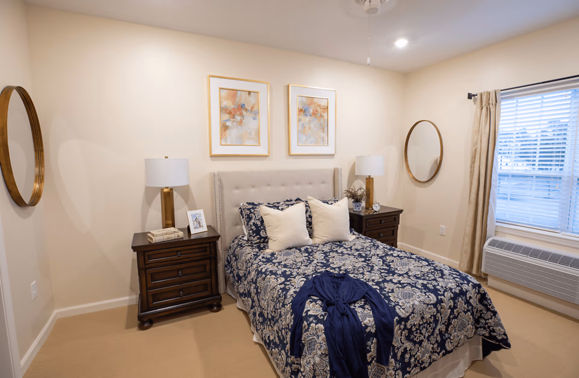 A neatly made bed with a blue and white patterned bedspread and two white pillows in a bedroom. There are two wooden nightstands on either side of the bed, each with a lamp and decorative items. Two abstract paintings hang above the bed. The room has beige walls, a window with blinds and beige curtains, a round mirror on each side wall, and a ceiling fan.