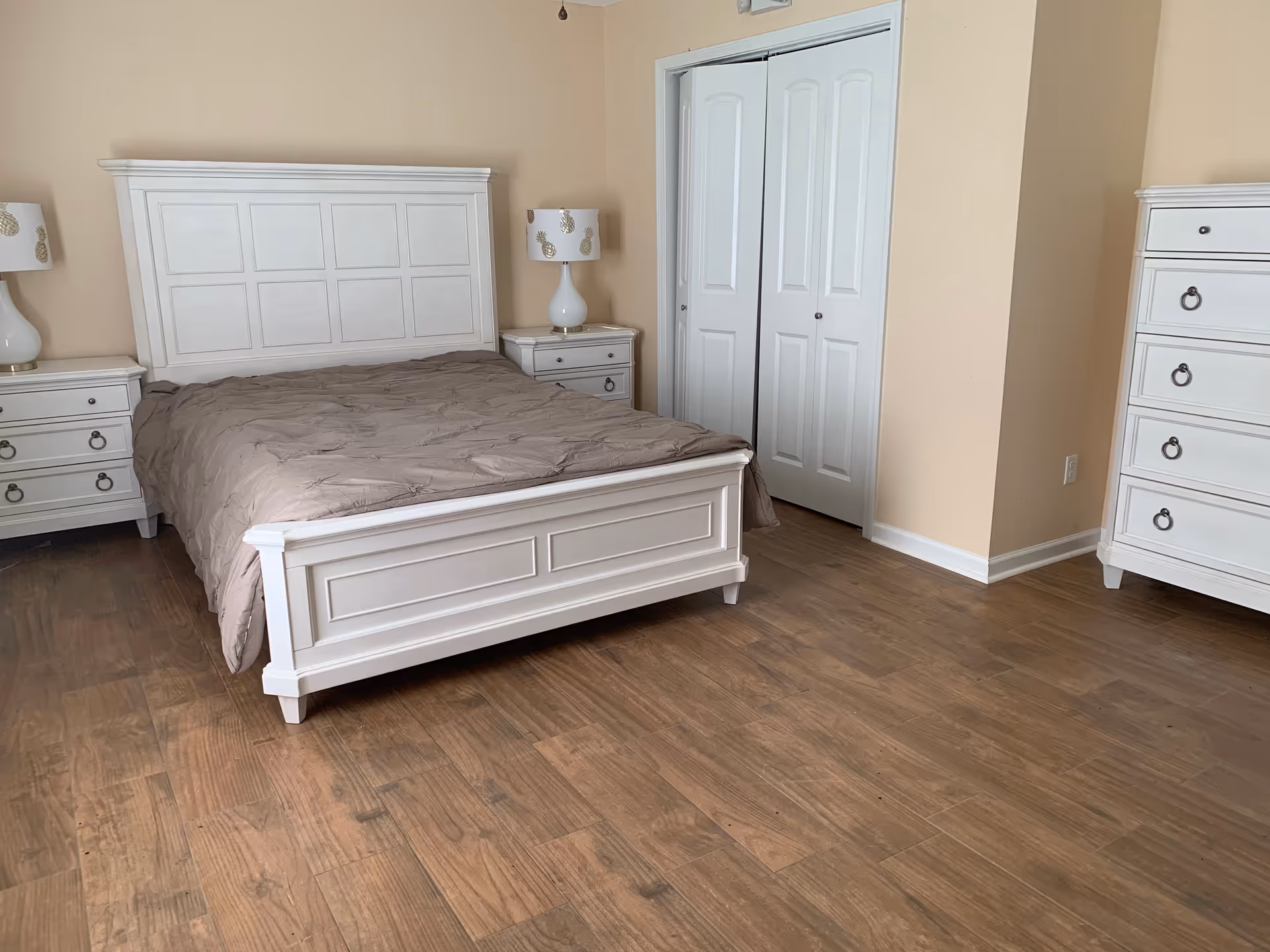A bedroom with a white wooden bed frame and matching white nightstands on either side, each with a lamp featuring a white shade with gold pineapples. There is a white dresser with drawers on the right side and a closet with white double doors in the background. The floor is wooden and the walls are painted beige.