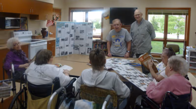 A group of elderly people, some in wheelchairs, gathered around a table in a room with kitchen cabinets and large windows. Two men stand at the far end of the table, one wearing a gray t-shirt and the other a gray button-up shirt. The table has photo boards and papers spread out on it.
