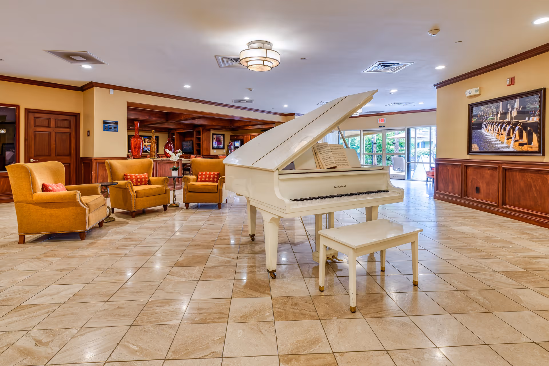 Spacious common area with a white grand piano and matching bench in the center. Surrounding the piano are four mustard-yellow armchairs with red patterned cushions. The room has beige tiled flooring, warm yellow walls with wooden paneling, and a large framed picture on the right wall. In the background, there are large glass doors leading outside and additional seating areas.