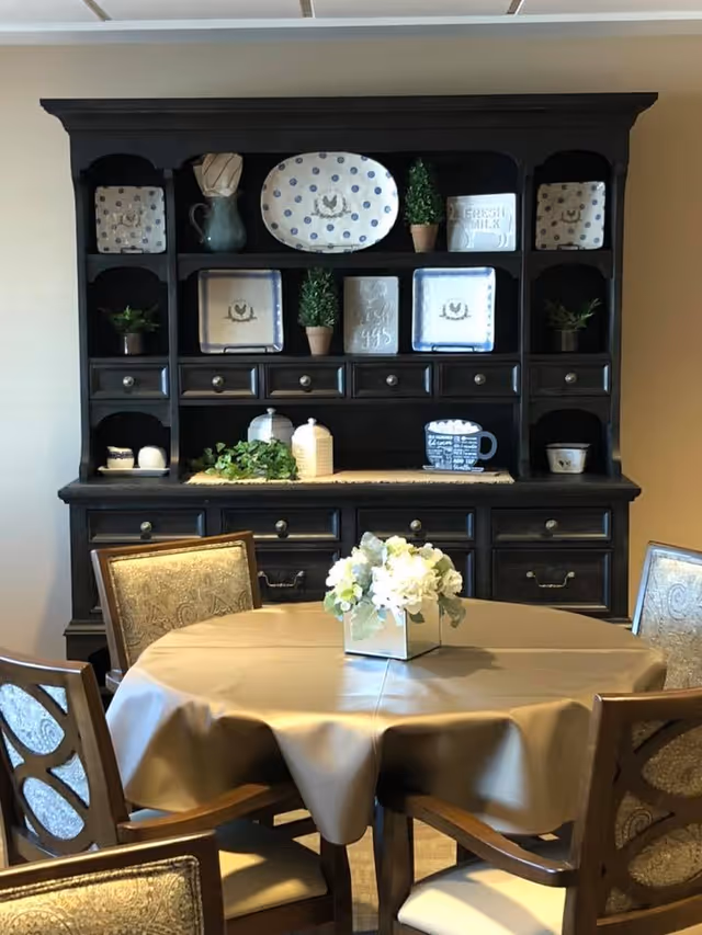 A dining area with a round table covered by a beige tablecloth and a centerpiece of white flowers in a square vase. Four wooden chairs with patterned upholstery surround the table. Behind the table is a large dark wooden hutch with multiple shelves and drawers, decorated with plates, small potted plants, and various decorative items.