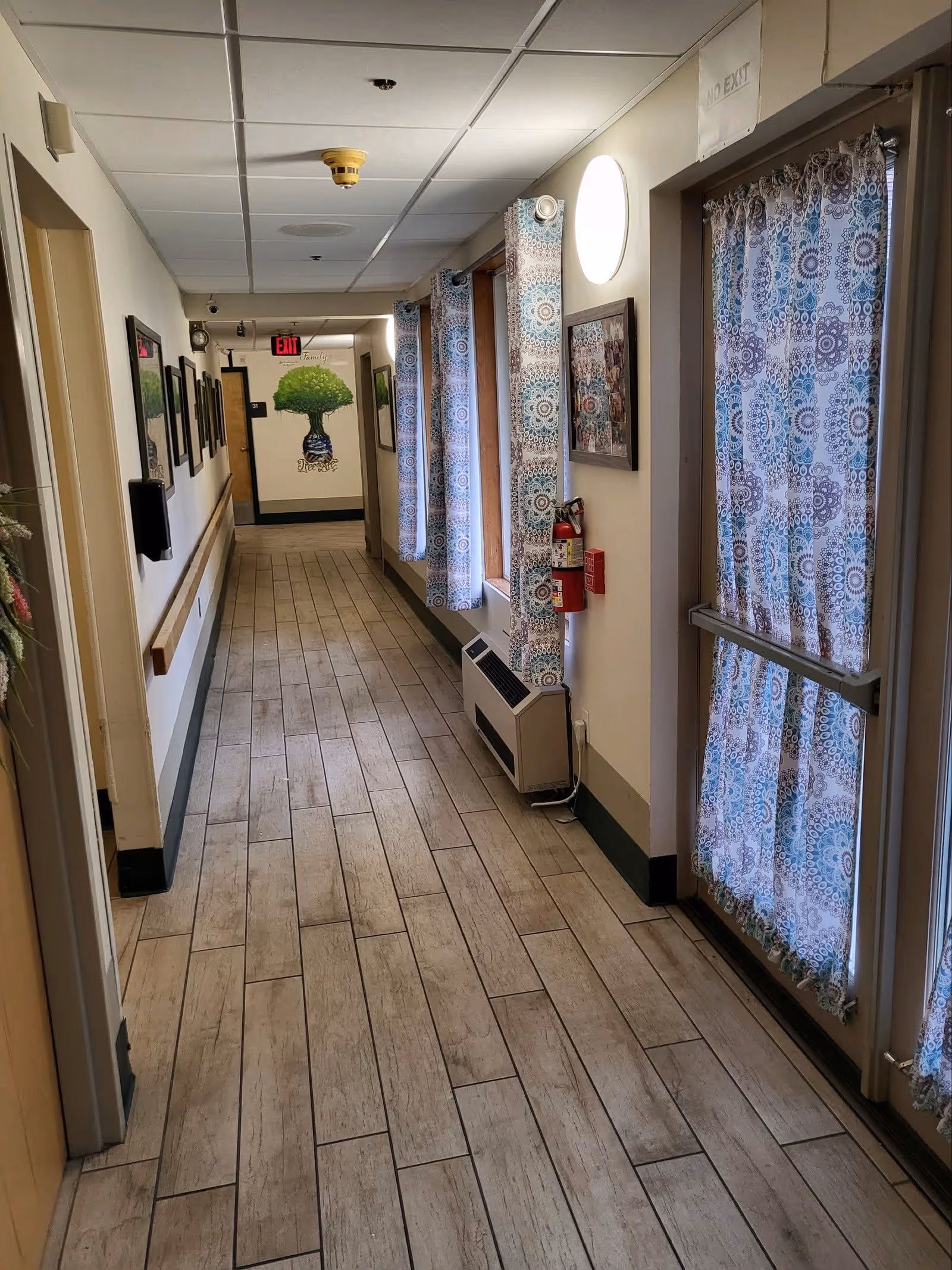 A well-lit hallway in a senior living facility with light wood-patterned tile flooring, white walls, and a drop ceiling. The right side features windows and a door covered with patterned blue and white curtains. There are framed pictures and a fire extinguisher mounted on the wall. At the end of the hallway, there is a mural of a tree with the words 'Family' and 'Memories' written above and below it. An exit sign is visible above a door at the end of the hallway.