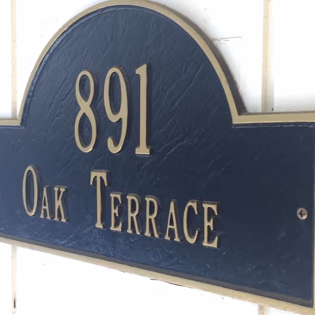 A close-up view of a dark blue and gold house number plaque mounted on a white wall, displaying the number 891 and the street name Oak Terrace.
