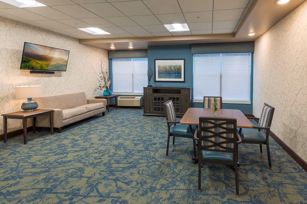 Well-lit communal living room with a sofa, wall-mounted TV, and a dining table with four chairs.