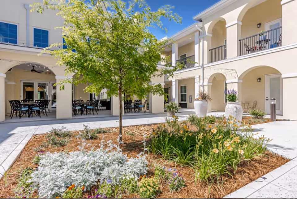 Outdoor courtyard area at The Blake at Panama City Beach featuring a landscaped garden with various plants and flowers, a small tree in the center, surrounded by a two-story building with arches and balconies. Several tables and chairs are placed under the covered patio area.