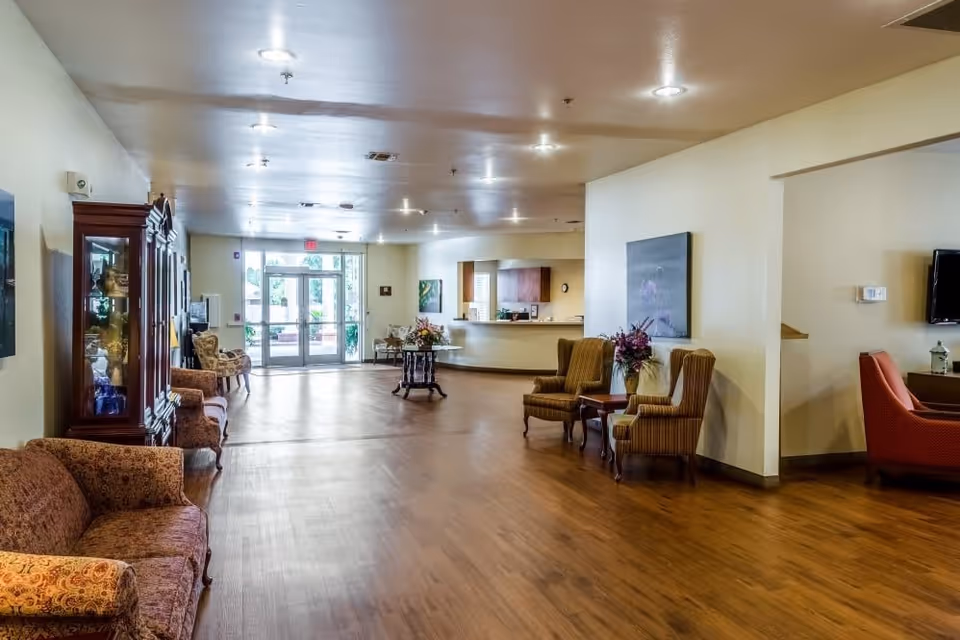 A spacious senior living facility common area with wooden flooring, multiple upholstered chairs and sofas, a wooden display cabinet, a small table with a flower arrangement, and a reception desk near the entrance with glass doors letting in natural light.