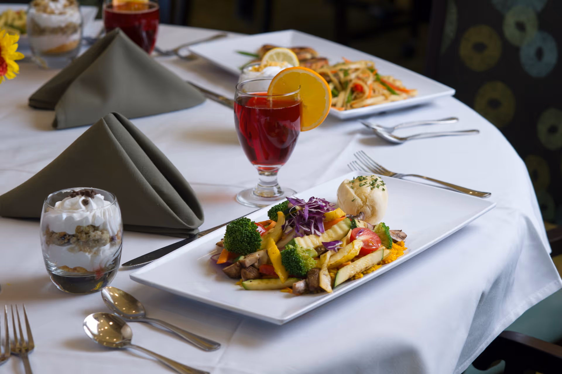 Plated entree of mixed vegetables with a roll, a dessert parfait, and a red beverage garnished with an orange slice on a white-tablecloth dining table.