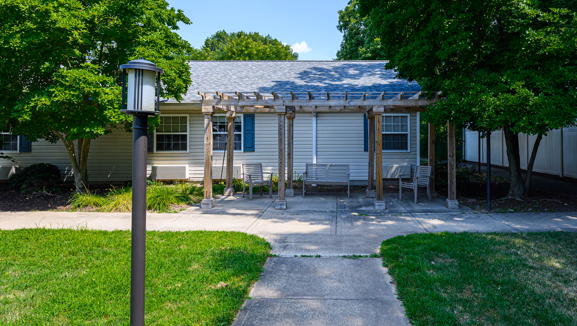 Outdoor seating area with wooden pergola and benches in front of a single-story building with white siding and blue shutters, surrounded by green trees and grass under a clear blue sky.