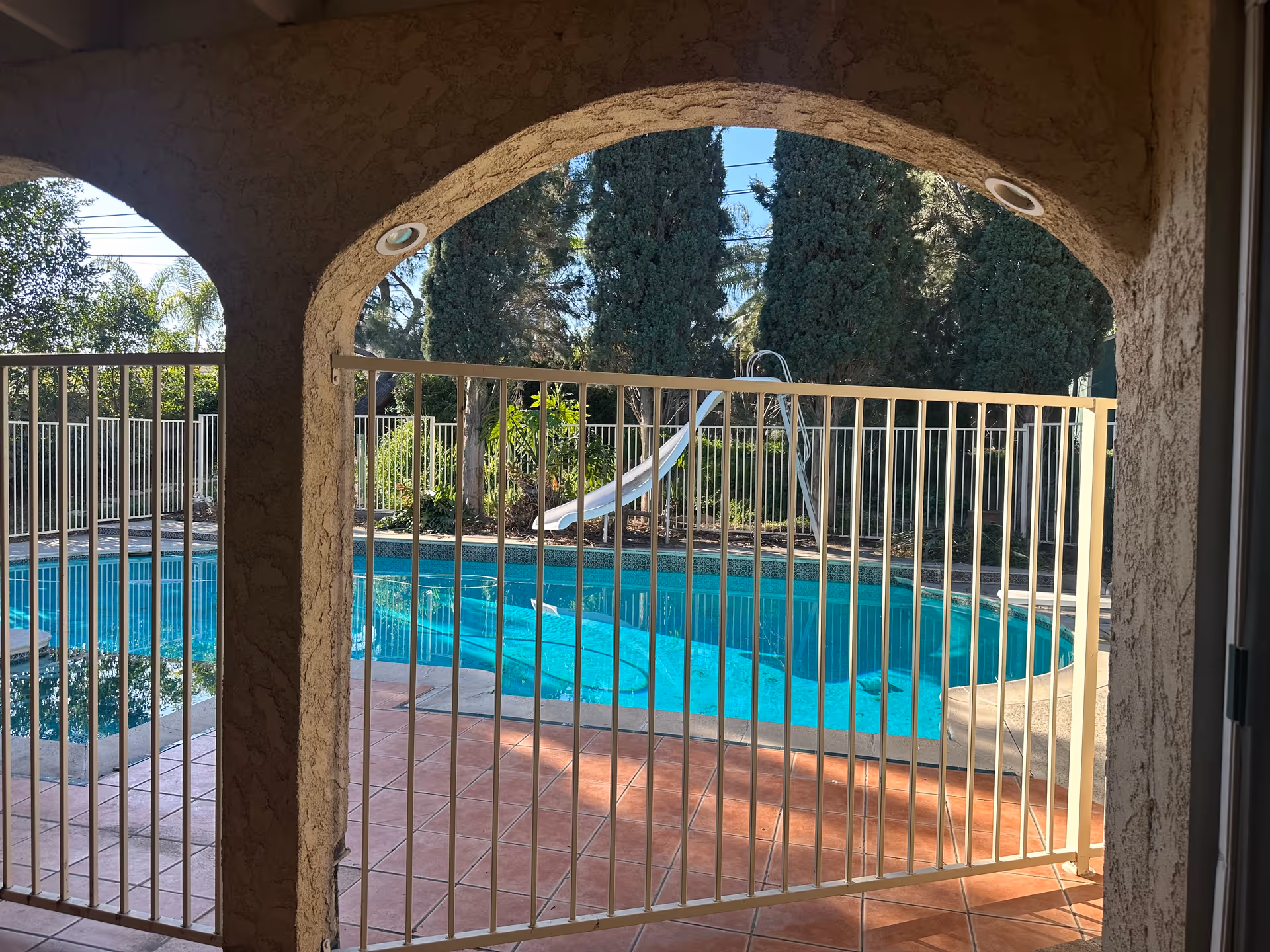 View of a swimming pool with a slide, surrounded by a safety fence, seen through an arched patio opening with textured walls. Trees and greenery are visible in the background under a clear blue sky.