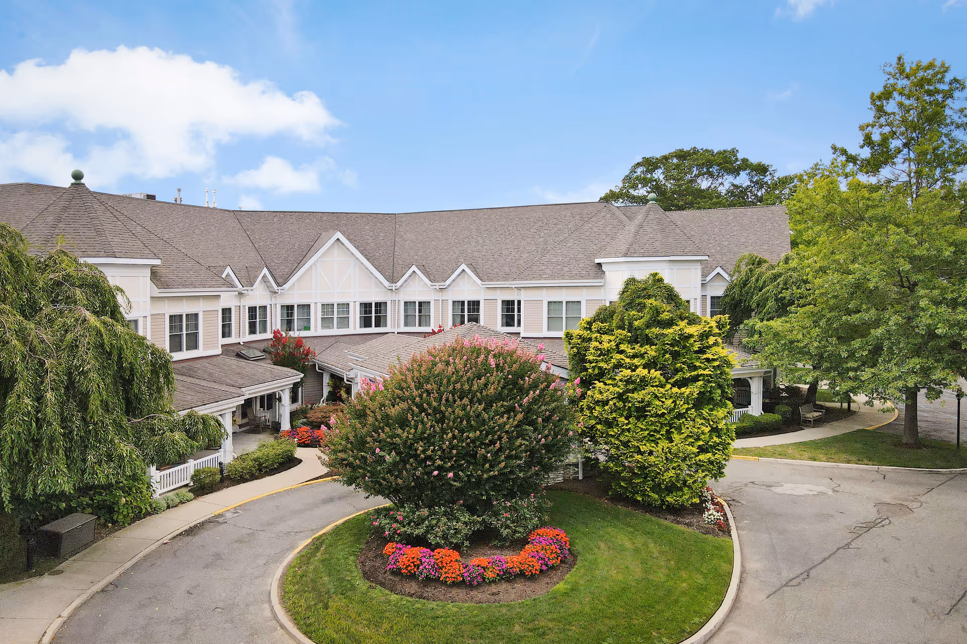 Exterior view of Sunrise of North Lynbrook senior living facility showing a large, two-story building with a gray roof and white trim. The building is surrounded by well-maintained landscaping including green trees, a large flowering bush, and colorful flower beds. A curved driveway leads to the entrance under a covered porch.