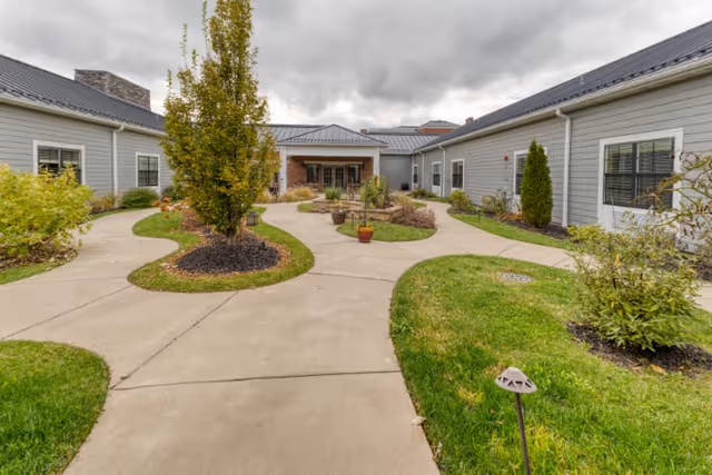 Outdoor courtyard area of a senior living facility with a concrete walkway curving around landscaped grass and plants, surrounded by single-story gray buildings under a cloudy sky.