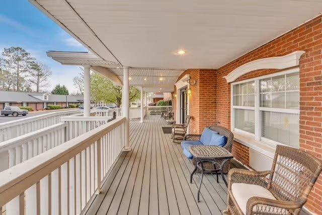 Covered outdoor porch area with wooden flooring and white railing, featuring wicker chairs with cushions and a small table, adjacent to a brick building with windows.