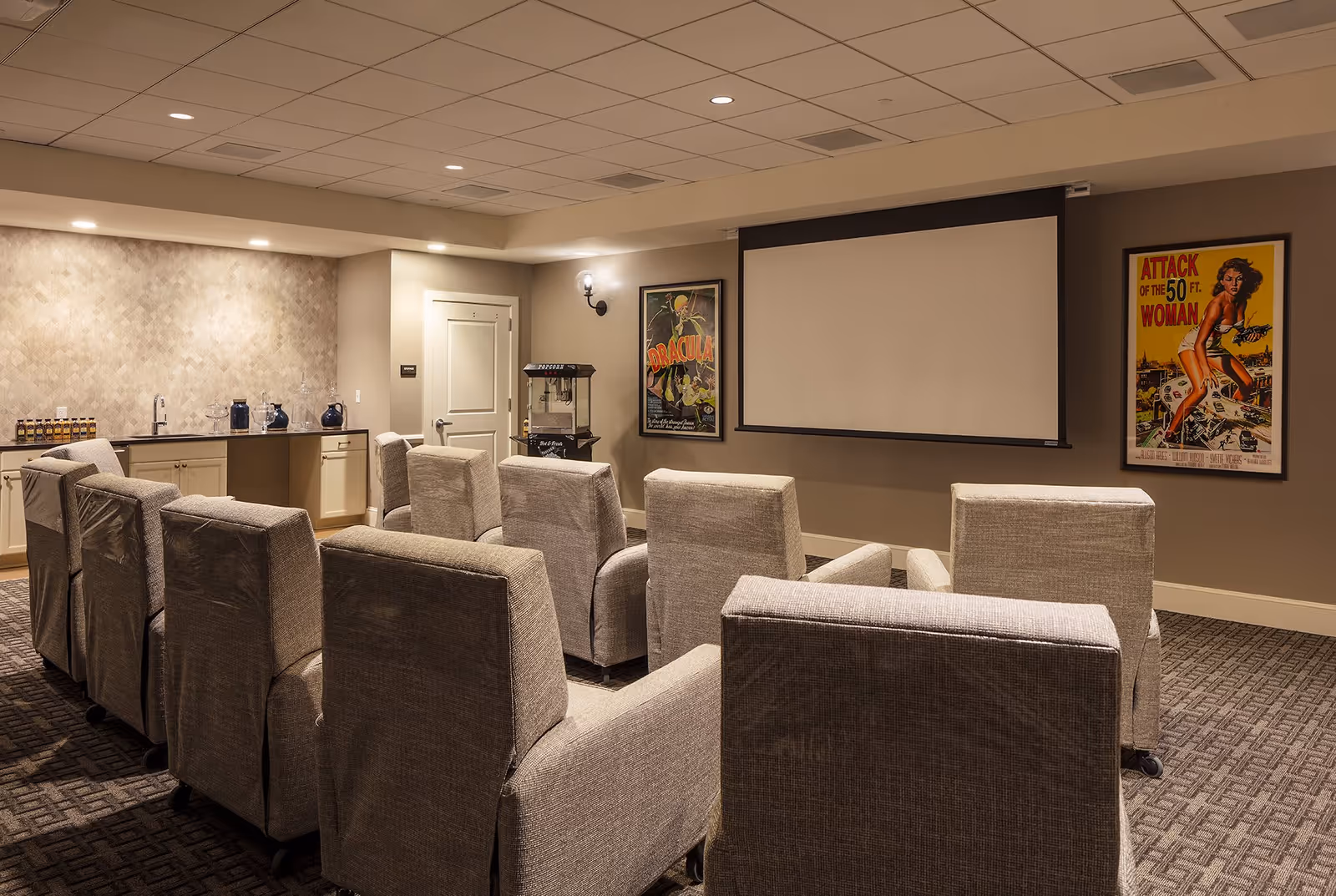 A cozy movie theater room with two rows of gray upholstered recliner chairs facing a large white projection screen. The walls are beige with two vintage movie posters, one for Dracula and one for Attack of the 50 Ft. Woman. There is a popcorn machine and a small counter with a sink and beverage dispensers on the left side of the room.