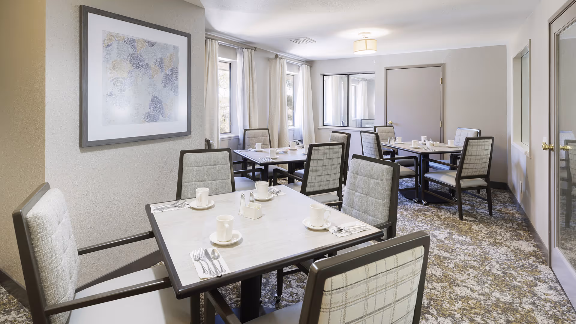 A dining room with several square tables, each set with white cups, saucers, and silverware. The room has cushioned chairs with patterned upholstery, light-colored walls, a framed abstract artwork, windows with curtains, and a carpeted floor.