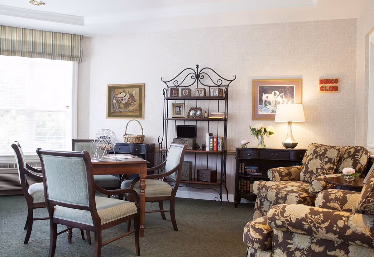 A cozy common room with a wooden table and four upholstered chairs arranged around it. On the table is a bingo cage and papers. Behind the table is a black metal shelving unit with framed photos, books, and decorative items. To the right, there are two floral patterned armchairs with a small round table between them holding a flower vase. A lamp on a black sideboard illuminates the room, which also features framed artwork on the walls and a window with striped valance letting in natural light. A small sign on the wall reads 'BINGO CLUB'.