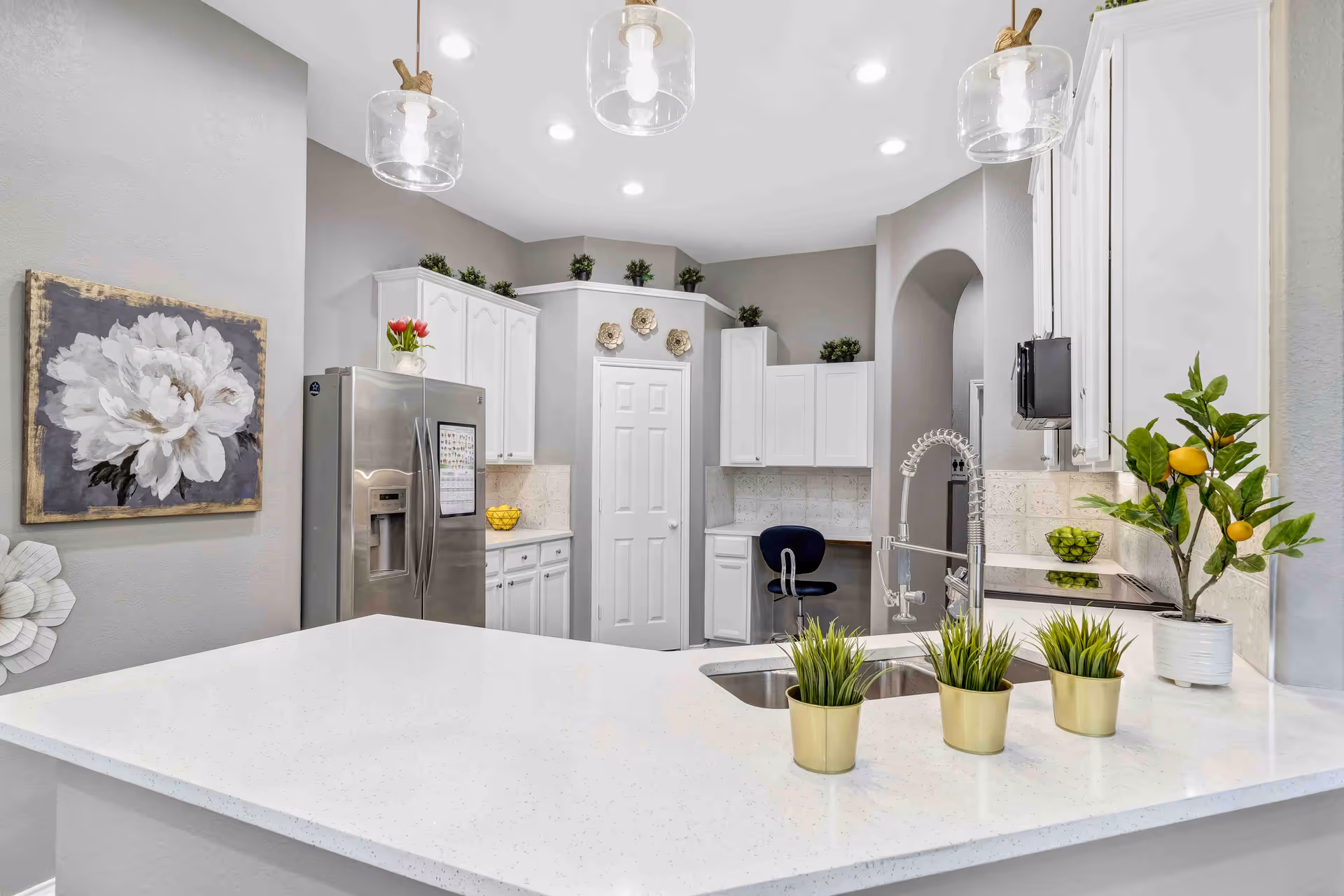A modern kitchen with white cabinets and countertops, a stainless steel refrigerator, a double sink with a flexible faucet, and three small potted plants on the countertop. The kitchen is decorated with a painting of a white flower, small green plants on top of the cabinets, and a lemon tree plant on the counter. There is a black chair tucked under a small desk area in the corner.