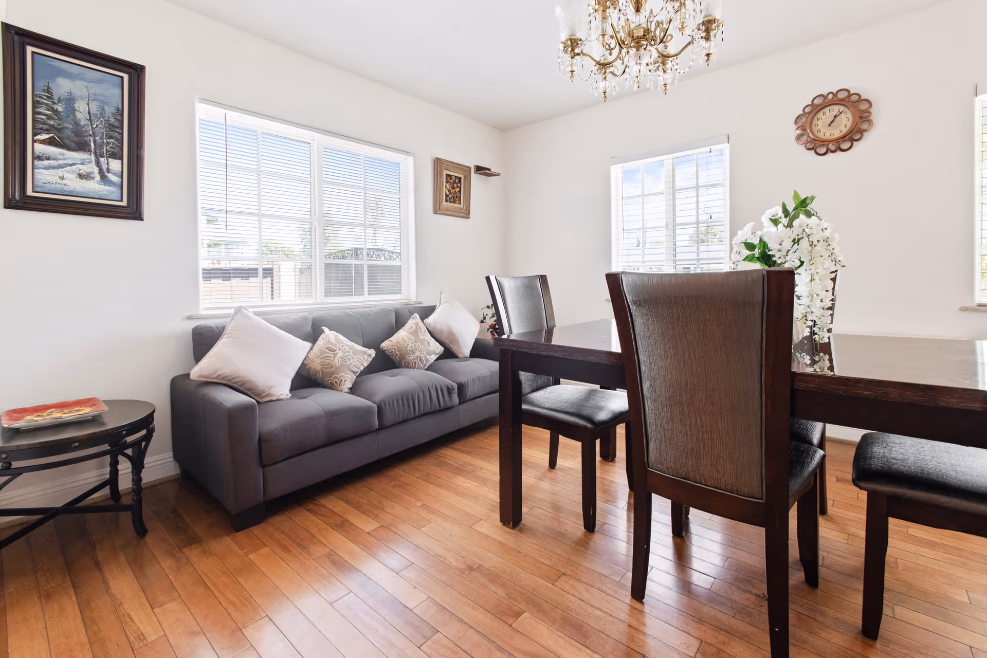 Bright combined living and dining room with a gray sofa, wooden dining table and chairs on hardwood floors.