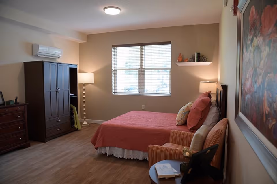 A cozy bedroom with a bed covered in a red quilt and multiple pillows, a striped armchair with a cushion, a small round table with books and a photo frame, a tall dark wooden wardrobe, a dresser, a floor lamp, a window with blinds, and a wall-mounted shelf with decorative items.