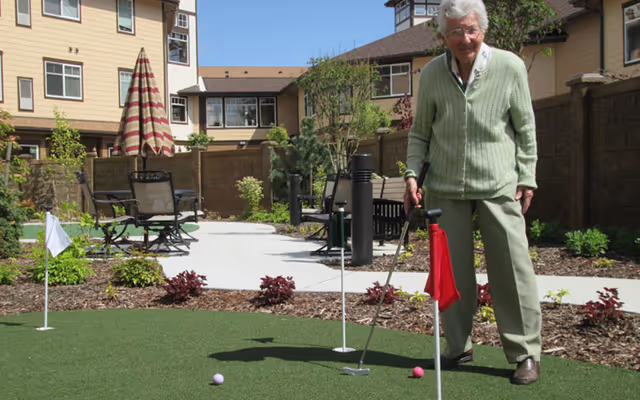 An elderly woman playing mini golf on a putting green in an outdoor courtyard area of a senior living facility. The courtyard has patio chairs, tables, plants, and surrounding buildings under a clear blue sky.